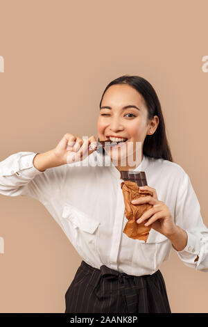 Freestyle. Jeune femme en shirt isolés sur bage mordre une barre de chocolat à l'appareil photo avec un clin d'enjoué Banque D'Images