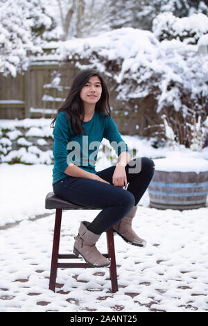 Teen girl relaxing Biracial sur tabouret à l'extérieur en hiver avec cour couverte de neige en arrière-plan Banque D'Images