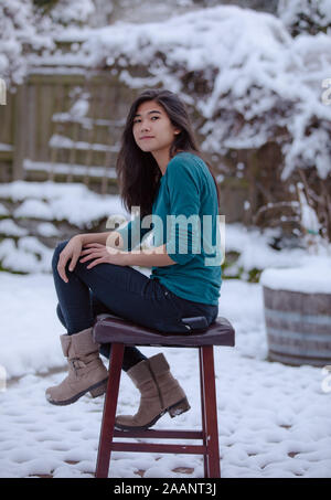 Teen girl relaxing Biracial sur tabouret à l'extérieur en hiver avec cour couverte de neige en arrière-plan Banque D'Images