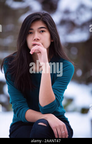 Teen girl relaxing Biracial sur tabouret à l'extérieur en hiver avec cour couverte de neige en arrière-plan Banque D'Images