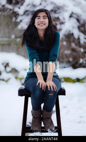 Teen girl relaxing Biracial sur tabouret à l'extérieur en hiver avec cour couverte de neige en arrière-plan Banque D'Images