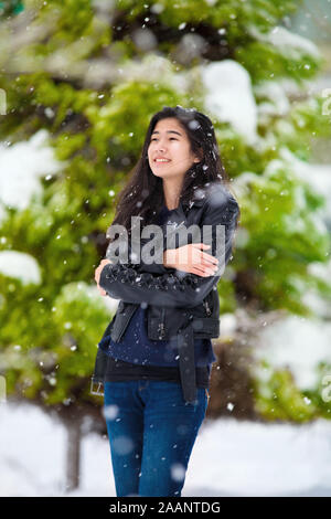 Jeune fille de l'adolescence biracial en jeans et blouson de cuir noir à l'extérieur froid frissons permanent en hiver avec des chutes de neige importantes. Je pine tree couvertes de neige Banque D'Images