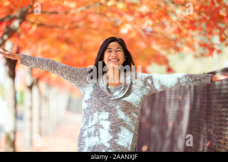 Thai Biracial American teen girl dans chandail gris et rouge vif, sous l'érable feuille orange à l'automne, avec les bras tendus, souriant pis Banque D'Images