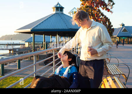 Woman en quarantaine biracial alimentation fils handicapé en fauteuil roulant à park sur sunny day Banque D'Images