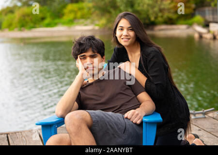 Frères et sœurs adolescents Biracial relaxant en plein air en bleu chaises Adirondack par Lake sur la jetée en bois Banque D'Images