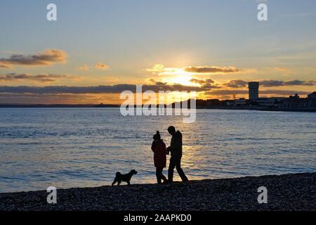 Silhouette d'un couple en train de marcher un chien sur la plage de Portsmouth dans un contexte d'un coucher de soleil d'hiver, Hampshire England UK Banque D'Images