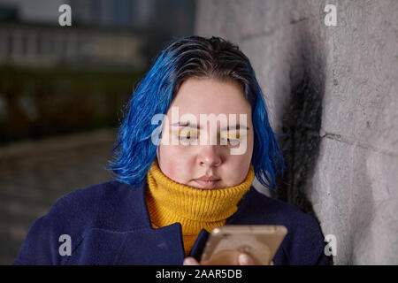 Une jeune femme rondelette se lit les messages sur un écran lors d'une froide soirée d'automne au crépuscule. Une fille avec les cheveux teints en bleu et un manteau bleu foncé utilise un smartph Banque D'Images