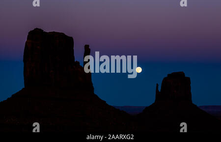 Pleine lune passant entre les silhouettes de l'emblématique mitten formations de Monument Valley après la tombée de la nuit. Banque D'Images