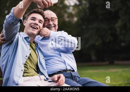 Man et petit-fils assis ensemble sur un banc de parc en tenant avec smartphone selfies Banque D'Images