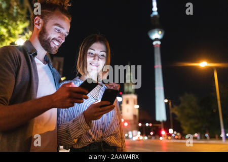 Couple heureux à l'aide de smartphones dans la ville la nuit, Fernsehturm en arrière-plan, Berlin, Allemagne Banque D'Images
