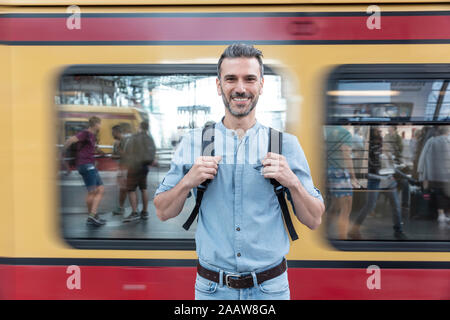 Portrait d'homme souriant à la plate-forme de la gare avec train en arrière-plan, Berlin, Allemagne Banque D'Images