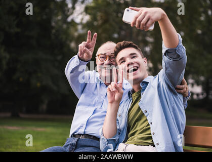 Man et petit-fils assis ensemble sur un banc de parc en tenant avec smartphone selfies Banque D'Images