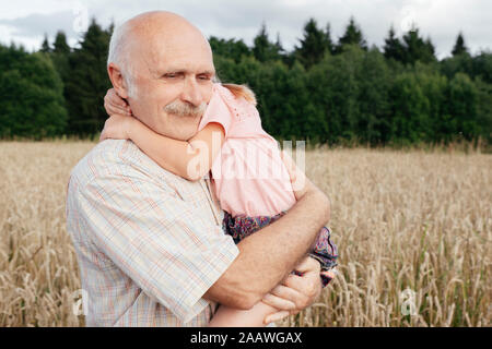 Portrait of senior man dans un champ d'avoine transportant sa petite-fille dans ses bras Banque D'Images