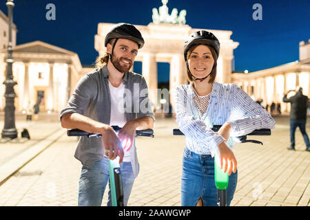 Jeune couple avec des scooters électriques à la porte de Brandebourg la nuit, Berlin, Allemagne Banque D'Images