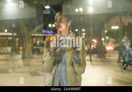 Portrait of smiling young woman with smartphone et regarder quelque chose d'écouteurs, Lisbonne, Portugal Banque D'Images
