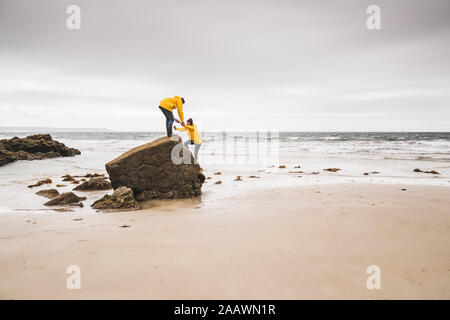 Jeune femme portant des vestes de pluie jaune et de l'escalade sur un rocher sur la plage, Bretagne, France Banque D'Images