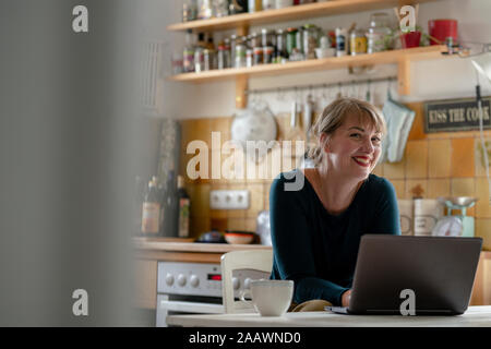 Portrait of smiling woman sitting in the kitchen using laptop Banque D'Images