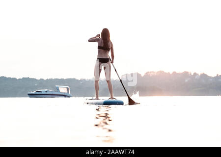 Vue arrière de jeune femme en stand up paddle à l'embarquement au coucher du soleil, le Lac de Starnberg, Allemagne Banque D'Images