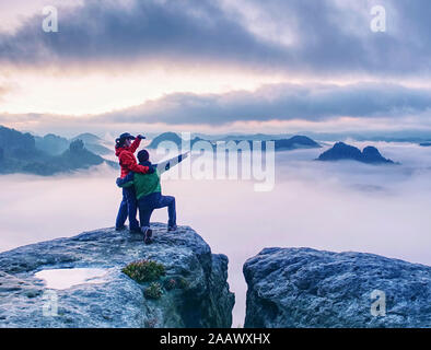 Les randonneurs la femme et l'homme dans les vêtements sombres sur plus haute montagne et regarder le lever du soleil dans l'obscurité. Sur le bord du sentier rocheux au-dessus de la vallée brumeuse. Tourist couple enj Banque D'Images