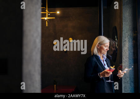 Femme d'âge mûr en prenant un cocktail après le travail, using smartphone Banque D'Images