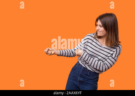 Vue de côté portrait de femme autoritaire avec les cheveux bruns en chemise rayée manches longues debout avec les poings serrés, faisant semblant de tirer avec beaucoup d'effort. Banque D'Images