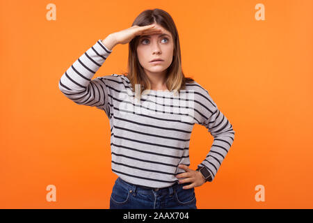 Portrait de jeune femme curieux concentré avec les cheveux bruns en chemise rayée manches longues standing looking loin avec la main au-dessus des yeux, surpris stare