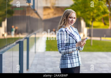 Portrait de belle businesslady élégant avec des cheveux blonds en veste et jupe à carreaux holding smartphone, à la confiance et succès, smiling at Banque D'Images