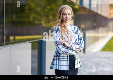 Portrait of attractive confiant businesslady avec de longs cheveux blonds en veste et jupe à carreaux debout avec les bras croisés et regarder avec trop de l'appareil photo Banque D'Images