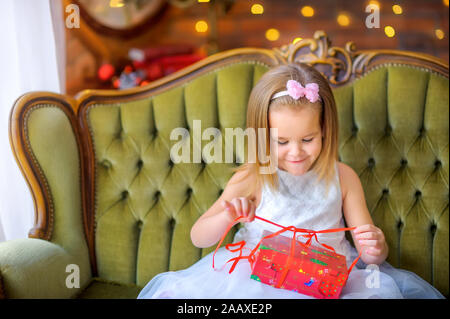 Happy little girl dans une robe est assise sur un canapé avec une boîte cadeau dans ses mains s'ouvre et sourit. Dénoue le ruban rouge. Noël et Nouvel An Banque D'Images