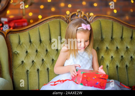 Happy little girl dans une robe est assise sur un canapé avec une boîte cadeau dans ses mains s'ouvre et sourit. Noël et Nouvel An Banque D'Images