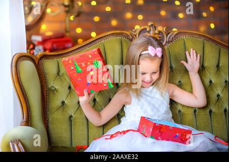 Happy little girl dans une robe est assise sur un canapé avec une boîte cadeau dans ses mains s'ouvre et sourit. Noël et Nouvel An Banque D'Images