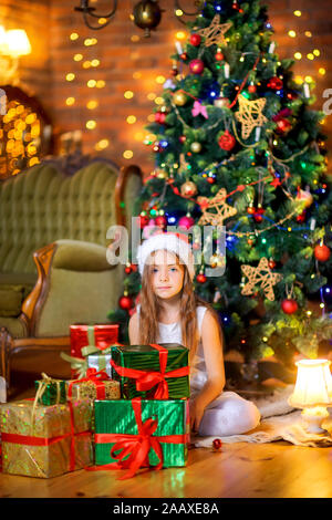 Une jolie jeune fille en robe blanche et Santa Claus hat est assis sur le plancher avec un gros tas de cadeaux. Dans le contexte d'un arbre de fête. Christm Banque D'Images