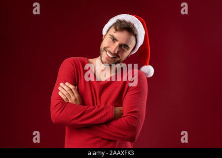 Beau jeune homme de race blanche en rouge chandail et Santa hats se dresse sur fond rouge en studio et des dents en souriant. Close up portrait of guy européenne avec C Banque D'Images