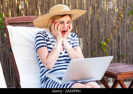 Oh mon Dieu ! Portrait de jeunes adultes réaction fou freelancer femme en robe et chapeau assis sur banquette-lit avec coffre, regardant avec de grands yeux d'un ordinateur Banque D'Images