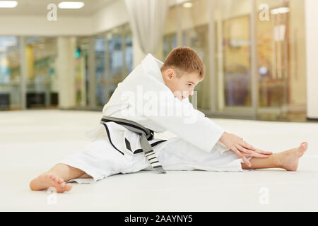 Little Boy wearing kimono blanc assis sur le plancher et l'échauffement avant l'entraînement en karaté il fait des exercices d'étirement Banque D'Images