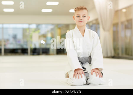 Portrait of little boy wearing kimono blanc assis sur le plancher et looking at camera il prêt pour l'entraînement sportif Banque D'Images