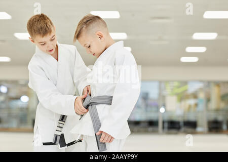 Garçon en kimono blanc montrant à son ami comment attacher la ceinture en ils debout dans la salle de sport Banque D'Images