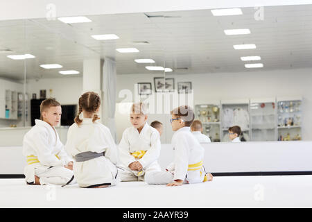 Groupe d'enfants en kimono assis sur le plancher et de parler les uns aux autres pendant leur leçon dans le karaté dans la salle de sport Banque D'Images