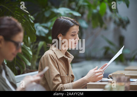 Side view portrait of young businesswoman reading documents tout en travaillant dans un café en plein air, copy space Banque D'Images