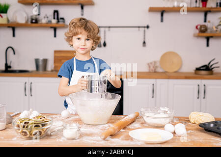 Cute little boy tamisant la farine sur bol en allant faire de la pâte de pâtisserie maison dans la cuisine Banque D'Images