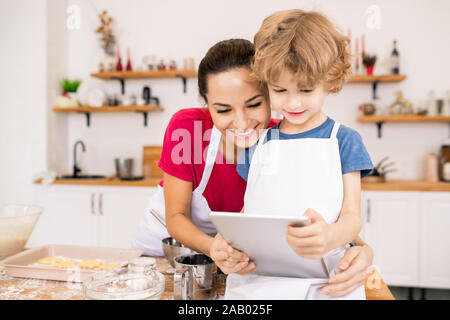 Tendre jeune femme et son fils avec surface tactile regardant la vidéo recette dans touchpad en allant faire cuire le dîner Banque D'Images