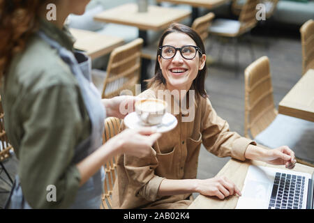 High angle portrait of young woman smiling at serveuse apportant une tasse à café tout en travaillant dans un café, copy space Banque D'Images