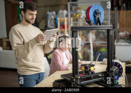 Jeune homme avec surface tactile à la recherche en ligne à la figure sur l'écran alors qu'il se tenait devant l'imprimante 3d sur fond de collègue Banque D'Images