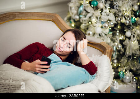 Young woman with mobile phone home allongé sur un canapé en face de sapin Banque D'Images