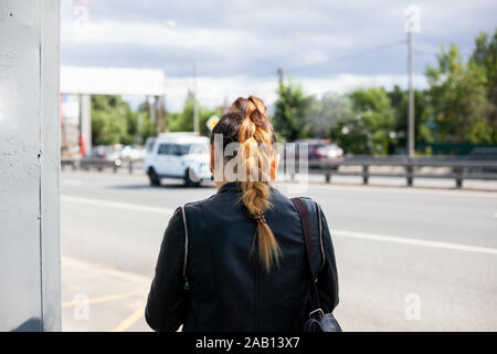 La jeune fille à l'arrêt de bus est en attente de l'autobus. Une femme se tient debout sur une route. Les cheveux teints. Un homme est en attente pour son transport. Personne inconnue sur le s Banque D'Images