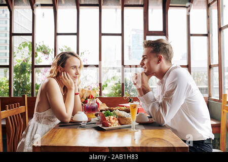 Smiling young man and woman sitting at table cafe avec beaucoup de plats délicieux et regarder avec amour et d'intérêt Banque D'Images