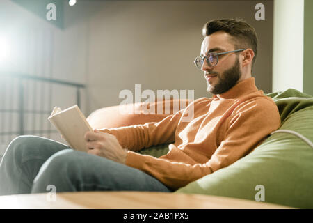 Jeune homme détendu dans casualwear et lunettes lecture livre sur un canapé confortable Banque D'Images