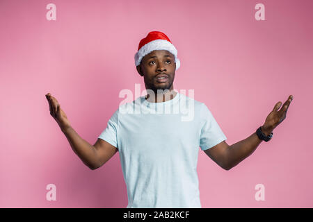 Jusqu'à la taille de balle déçu african american man étend les mains avec de l'indignation, wearing Santa hat, t shirt, jusqu'à la perplexité, isolé sur Rose Banque D'Images