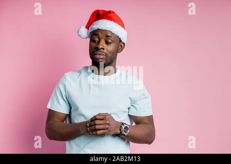 Coup horizontal teint foncé de jeune homme calme barbu in Santa hat, à côté avec les expression, gardant les mains ensemble, penser ou tr Banque D'Images