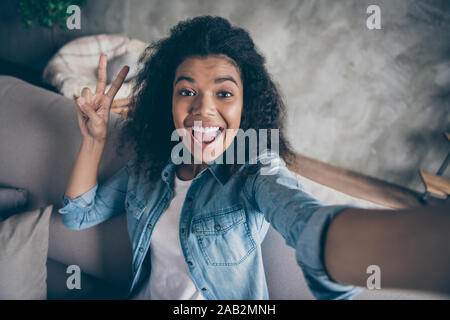 Photo gros plan de la peau sombre assez funky blogger curly lady faire excité autoportraits montrant v-sign symbole sitting Comfy Couch denim casual outfit vivant Banque D'Images